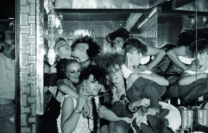 Group of young women in 1980s goth/punk fashion crowded into a mirrored bathroom at Foobert's nightclub in London, showing bold hair, makeup, and expressive camaraderie.