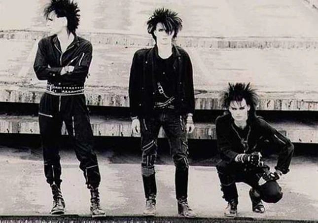 Members of Skinny Puppy in stylized dark clothing with spiked hair and DIY aesthetic, representing expressive masculine presence in industrial and alternative music scenes